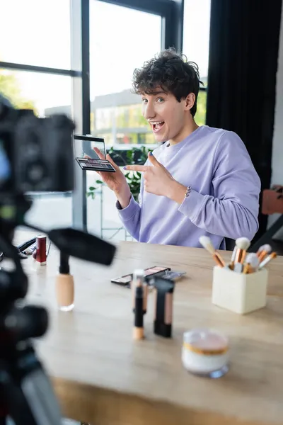 Excited transgender makeup artist pointing at eye shadows near cosmetics and digital camera — Stock Photo