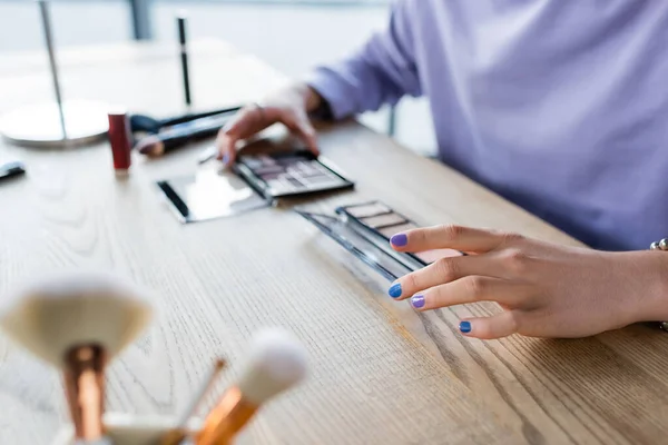Cropped view of transgender person holding eye shadows near blurred cosmetic brushes on table — Stock Photo