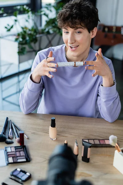 Smiling transgender person holding mascara near decorative cosmetics and blurred digital camera — Stock Photo