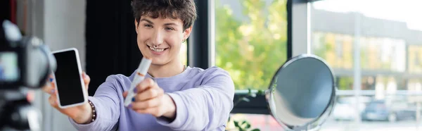 Une personne transgenre souriante tenant un mascara et un smartphone près d'un miroir flou et d'un appareil photo numérique, bannière — Photo de stock