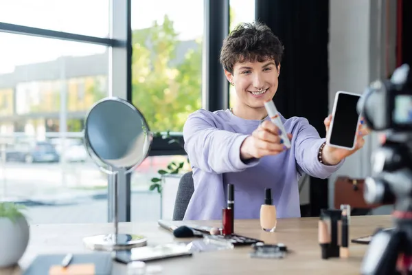 Sonriente persona transgénero sosteniendo rímel y teléfono inteligente cerca de cosméticos decorativos y cámara digital — Stock Photo