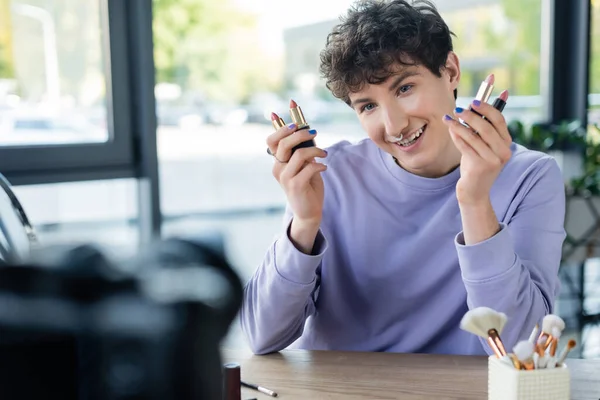 Positive transgender blogger holding lipsticks near blurred digital camera — Stock Photo