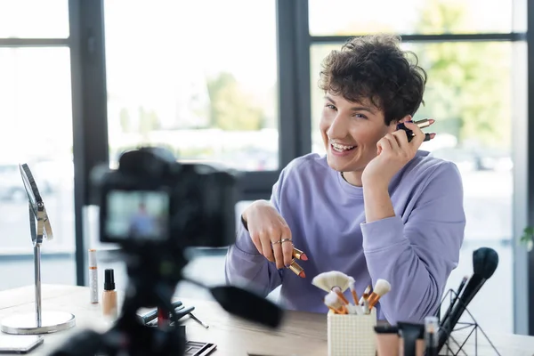 Young transgender person holding lipsticks near blurred digital camera and decorative cosmetics — Stock Photo