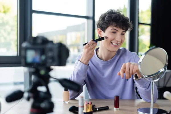 Positive transgender person applying face foundation near mirror and blurred digital camera — Stock Photo