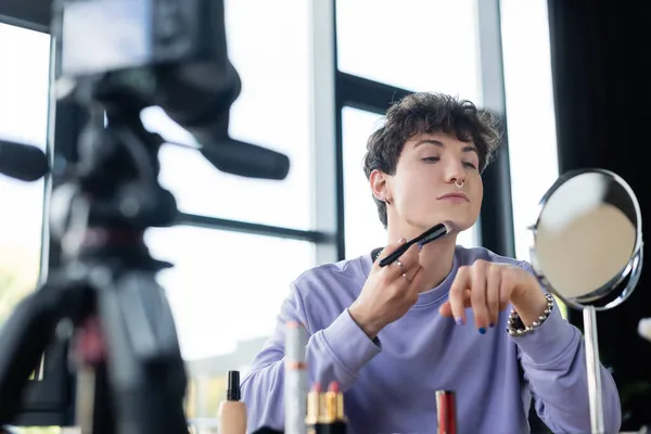 Jovem transgênero aplicando rosto fundação perto do espelho e câmera digital borrada — Fotografia de Stock