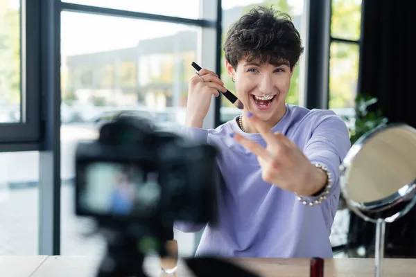 Positive transgender person holding cosmetic brush and showing peace sign near digital camera — Stock Photo