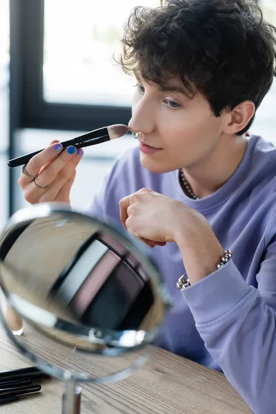 Jovem transgênero pessoa aplicando maquiagem fundação perto de espelho no estúdio — Fotografia de Stock