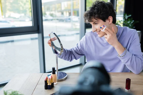Side view of smiling transgender person applying face powder ear mirror and blurred digital camera — Stock Photo
