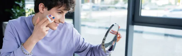 Vista laterale della persona transgender sorridente che applica polvere per il viso vicino allo specchio in studio, banner — Foto stock