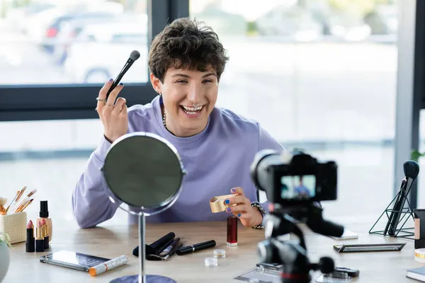 Cheerful transgender person holding face powder and cosmetic brush near digital camera and decorative cosmetics — Stock Photo