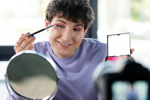 Giovane transgender persona che applica ombretto vicino specchio e fotocamera digitale offuscata — Foto stock