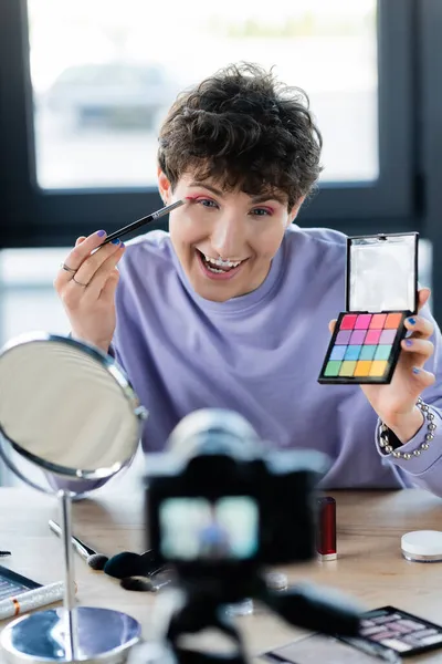 Excited transgender person applying eye shadow near mirror and digital camera — Stock Photo