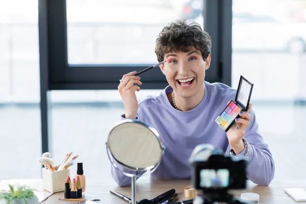 Happy transgender pessoa aplicando sombra nos olhos e olhando uma câmera perto de cosméticos decorativos e câmera digital — Fotografia de Stock