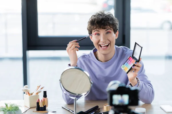Happy transgender person applying eye shadow and looking a camera near decorative cosmetics and digital camera — Stock Photo