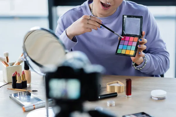 Vista cortada da pessoa transgênero segurando sombras de olhos perto de cosméticos e câmera digital — Fotografia de Stock