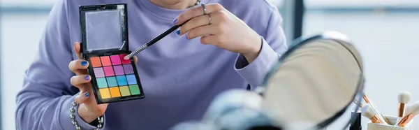 Cropped view of transgender person holding eye shadows and cosmetic brush near mirror, banner — Stock Photo