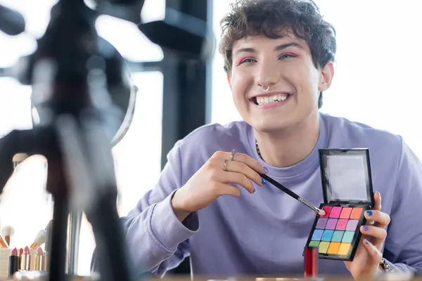 Smiling transgender person holding eye shadows and cosmetic brush near tripod and mirror — Stock Photo