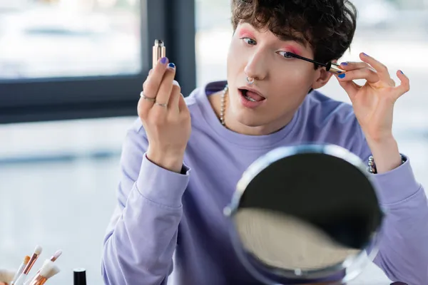 Persona transgender che applica mascara vicino pennelli cosmetici e specchio — Foto stock
