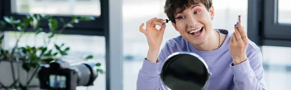 Sourire transgenre personne appliquant mascara près du miroir et appareil photo numérique, bannière — Photo de stock