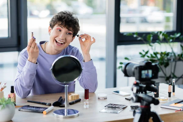 Persona transgender sorridente che applica il mascara vicino ai cosmetici decorativi e alla fotocamera digitale — Foto stock