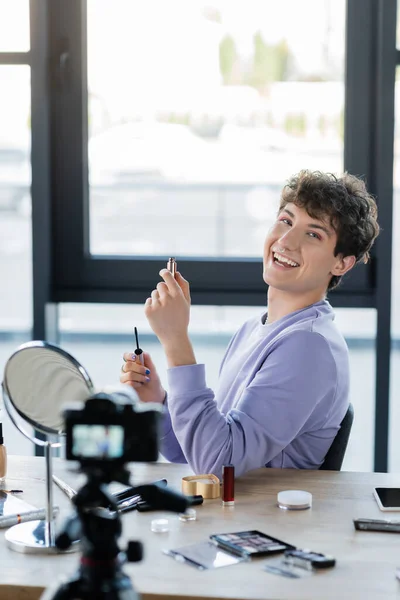 Positive transgender person holding mascara near smartphone, decorative cosmetics and digital camera — Stock Photo