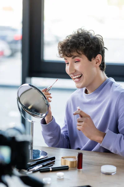 Cheerful transgender person holding lip balm near mirror and decorative cosmetics and digital camera — Stock Photo