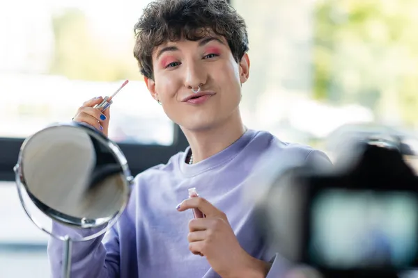 Transgénero persona sosteniendo bálsamo labial y pucheros labios cerca de espejo y cámara digital - foto de stock