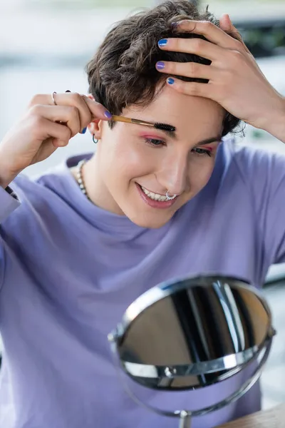 Sonriente persona transgénero cepillando cejas cerca del espejo - foto de stock
