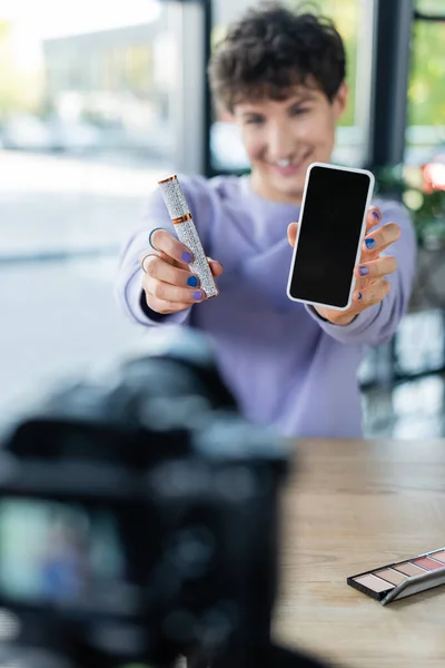 Rímel y smartphone en manos de una persona transgénero borrosa cerca de la cámara digital - foto de stock