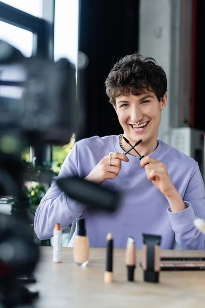 Sonriente persona transgénero sosteniendo cepillos de rímel cerca de cosméticos decorativos y cámara digital - foto de stock