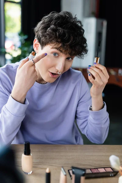 Transgender person holding mascara and pointing at eye near decorative cosmetics — Stock Photo
