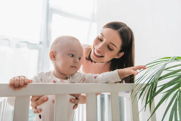 Happy mother holding baby boy touching plant while standing in crib — Stock Photo