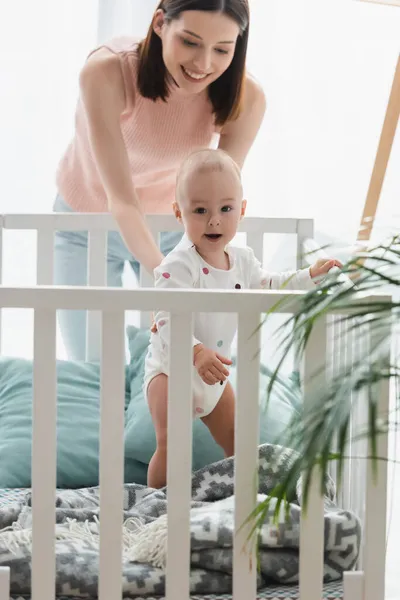 Little child in romper standing in crib near happy mother and blurred plant — Stock Photo