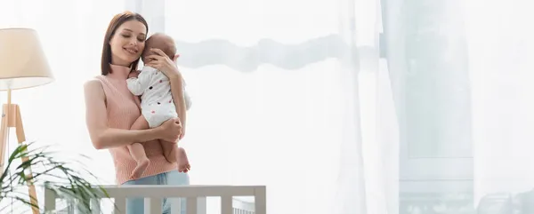 Happy woman with closed eyes hugging little son near crib at home, banner — Stock Photo