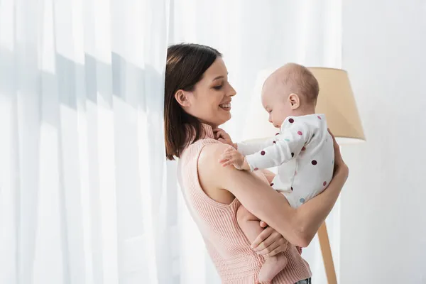 Side view of joyful woman holding little son near white curtain — Stock Photo