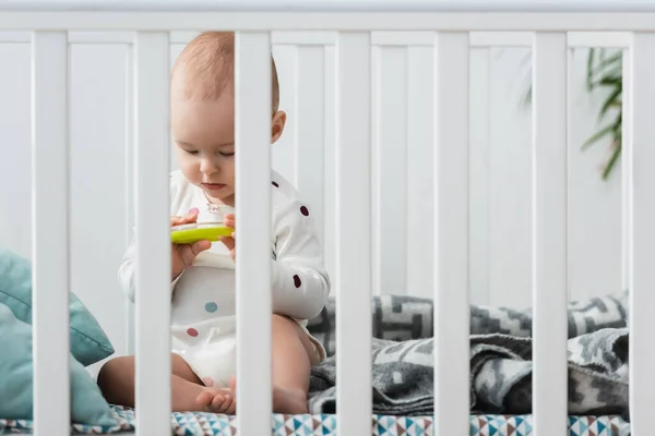 Niño pequeño sentado en la cuna y jugando con el anillo de sonajero - foto de stock