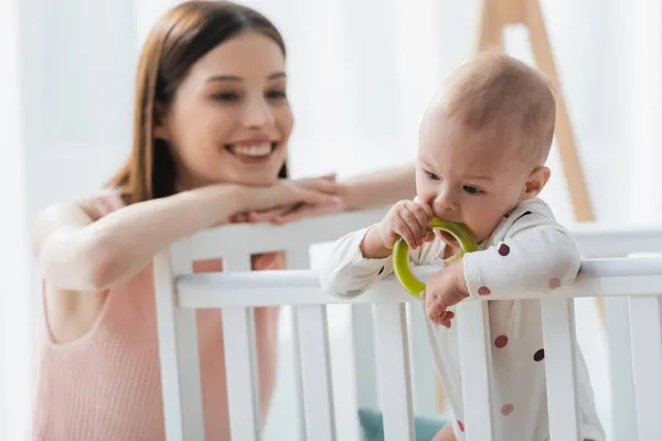 Blurred woman smiling while looking at little son biting rattle ring in crib — Stock Photo