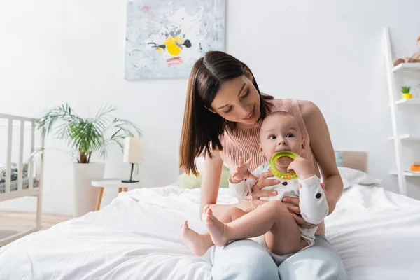 Smiling brunette woman sitting on bed with little son biting rattle ring — Stock Photo
