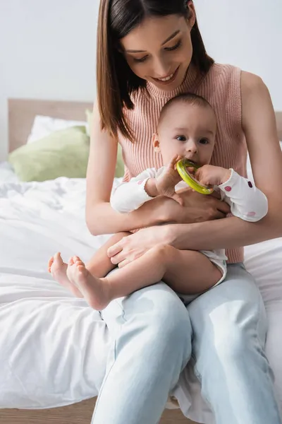 Smiling woman holding baby boy with rattle ring while sitting on bed — Stock Photo