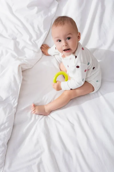 Над головой вид маленького мальчика с гремучим кольцом, смотрящего на камеру, сидя на белых кроватях — стоковое фото