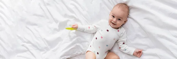 Top view of cheerful toddler boy lying on white bedding with rattle ring, banner — Stock Photo