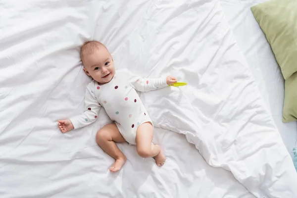 Top view of smiling baby boy with rattle ring looking at camera while lying on white bedding — Stock Photo