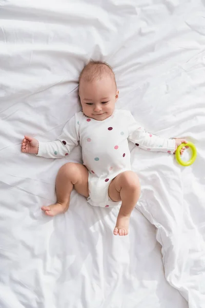 Top view of pleased child in romper lying on white bedding while holding rattle ring — Stock Photo