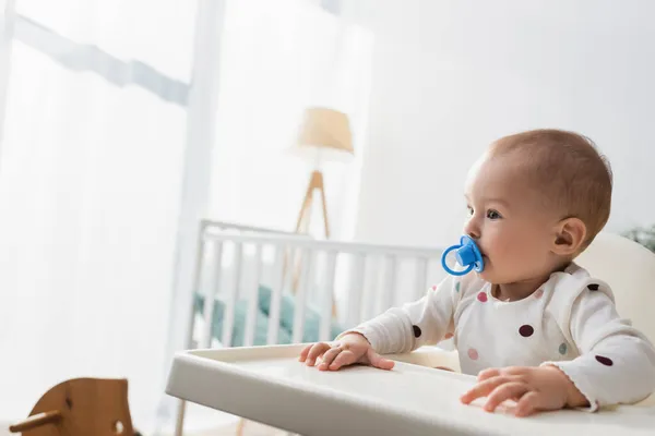 Tout-petit enfant avec sucette assis dans une chaise bébé près d'un berceau flou — Photo de stock