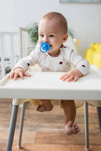 Glücklicher kleiner Junge mit Schnuller sitzt zu Hause im Kinderstuhl — Stockfoto