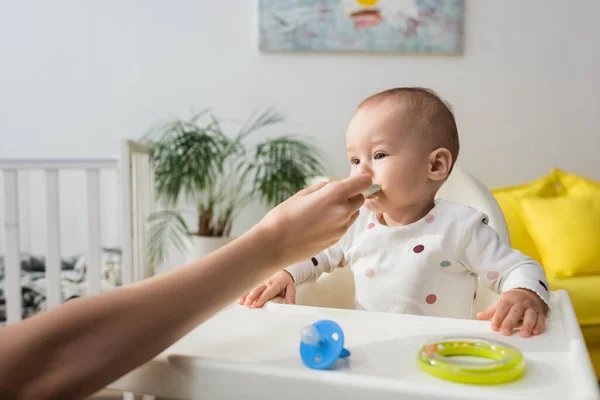 Mulher alimentando pequeno filho perto de chupeta e chocalho anel na cadeira do bebê — Fotografia de Stock