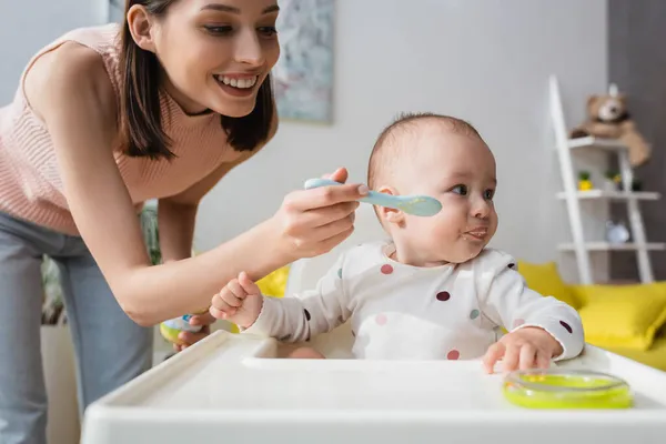 Sorridente donna bruna che nutre il piccolo figlio seduto sulla sedia del bambino — Foto stock