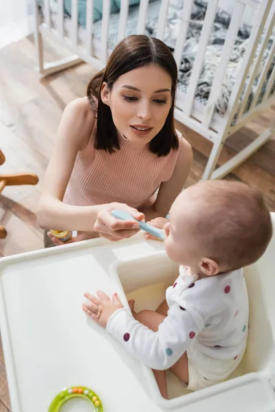 Vista ad alto angolo della donna bruna che alimenta il figlio offuscato sulla sedia del bambino — Foto stock