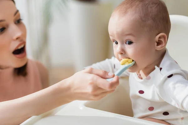 Tout-petit enfant en chaise bébé manger de la purée près de mère floue — Photo de stock