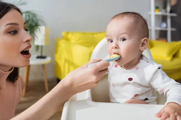 Brunette woman with open mouth feeding toddler kid in baby chair — Stock Photo